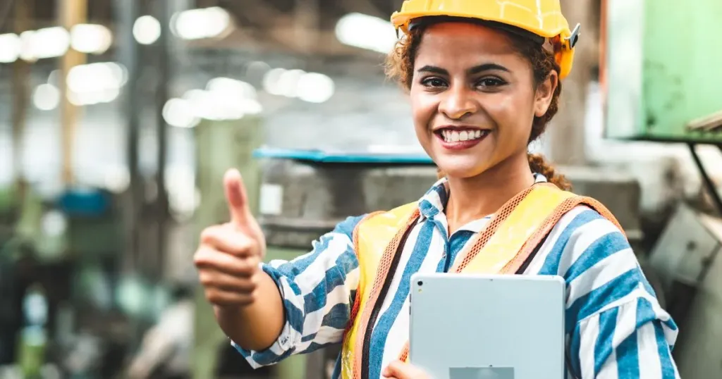 Industrial engineer wearing a hard hat and safety vest, holding a tablet on the shop floor, with machines in the background
