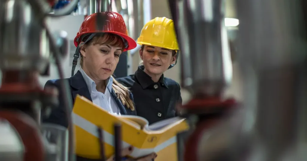 Two professionals wearing hard hats check a technical document among the piping of an industrial facility