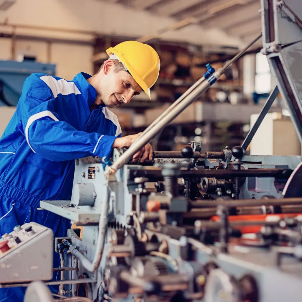 Technician in a hard hat and blue coveralls performing maintenance on an industrial machine with shafts and gears