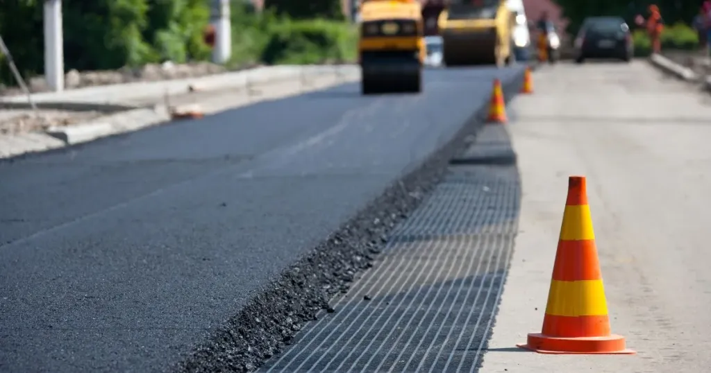 Road roller compacting asphalt mix on a road, with a reinforcement grid laid over the base beside it and traffic cones